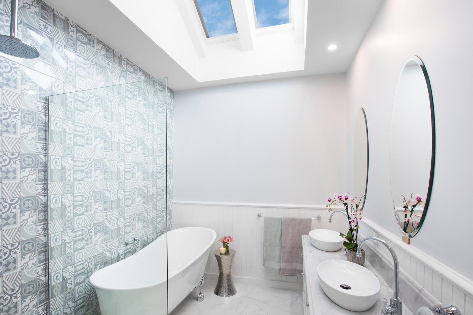 Bright bathroom with twin skylights, patterned feature tiles, a freestanding bathtub, glass shower screen and twin vessel sinks in a modern layout.