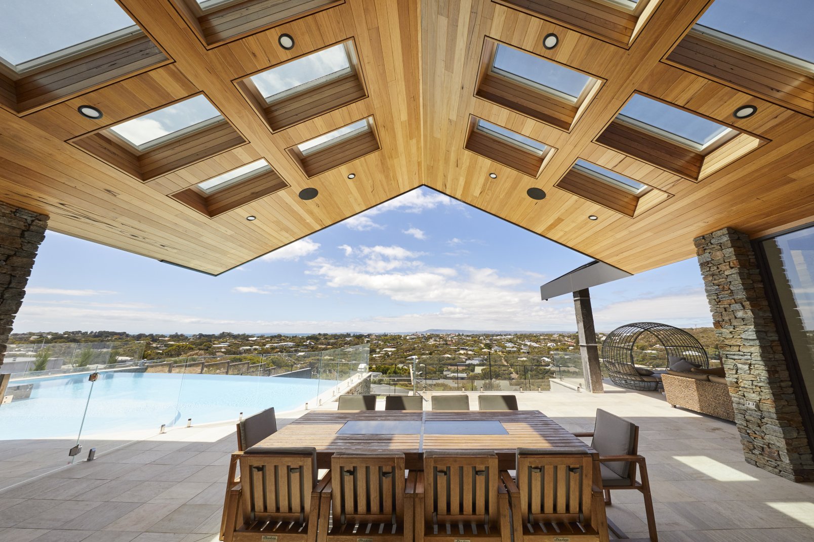 Timber patio ceiling with multiple skylights opens over an outdoor dining area, an infinity pool and wide coastal views on a bright, clear day.