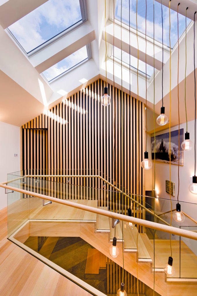 Bright modern stairwell with timber slat wall, skylights, glass balustrade, and hanging pendant lights over wooden stairs