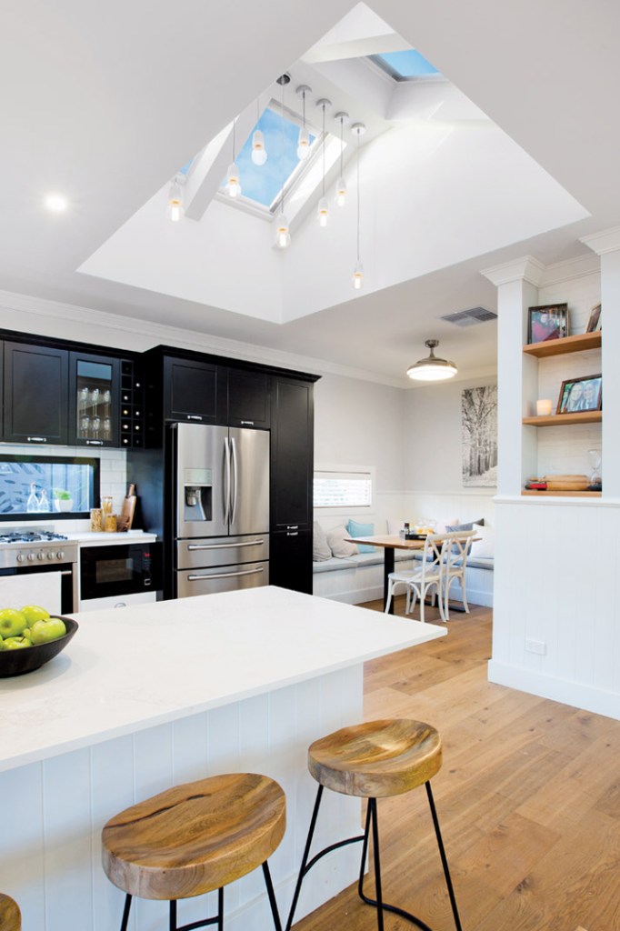Bright modern kitchen with skylights, black cabinetry, a white island, wooden stools, dining area, and indoor shelving